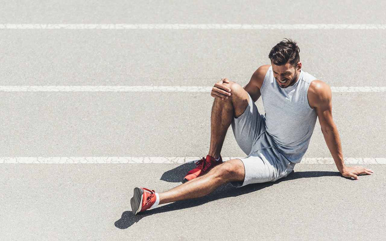 Knieschmerzen beim Laufen, Mann sitzt mit schmerzverzerrtem Gesicht auf einer grauen Tartanbahn